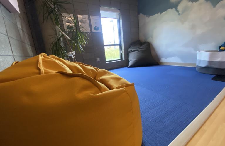 A beanbag in a room with a cloud mural and blue rug, as well as window letting in natural light