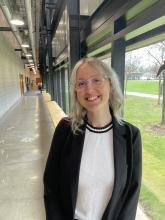Nicole is wearing a white top with a black blazer. She is standing in a hallway with a white wall on one side and glass windows on the other. You can see grass and some trees when looking out from the glass.